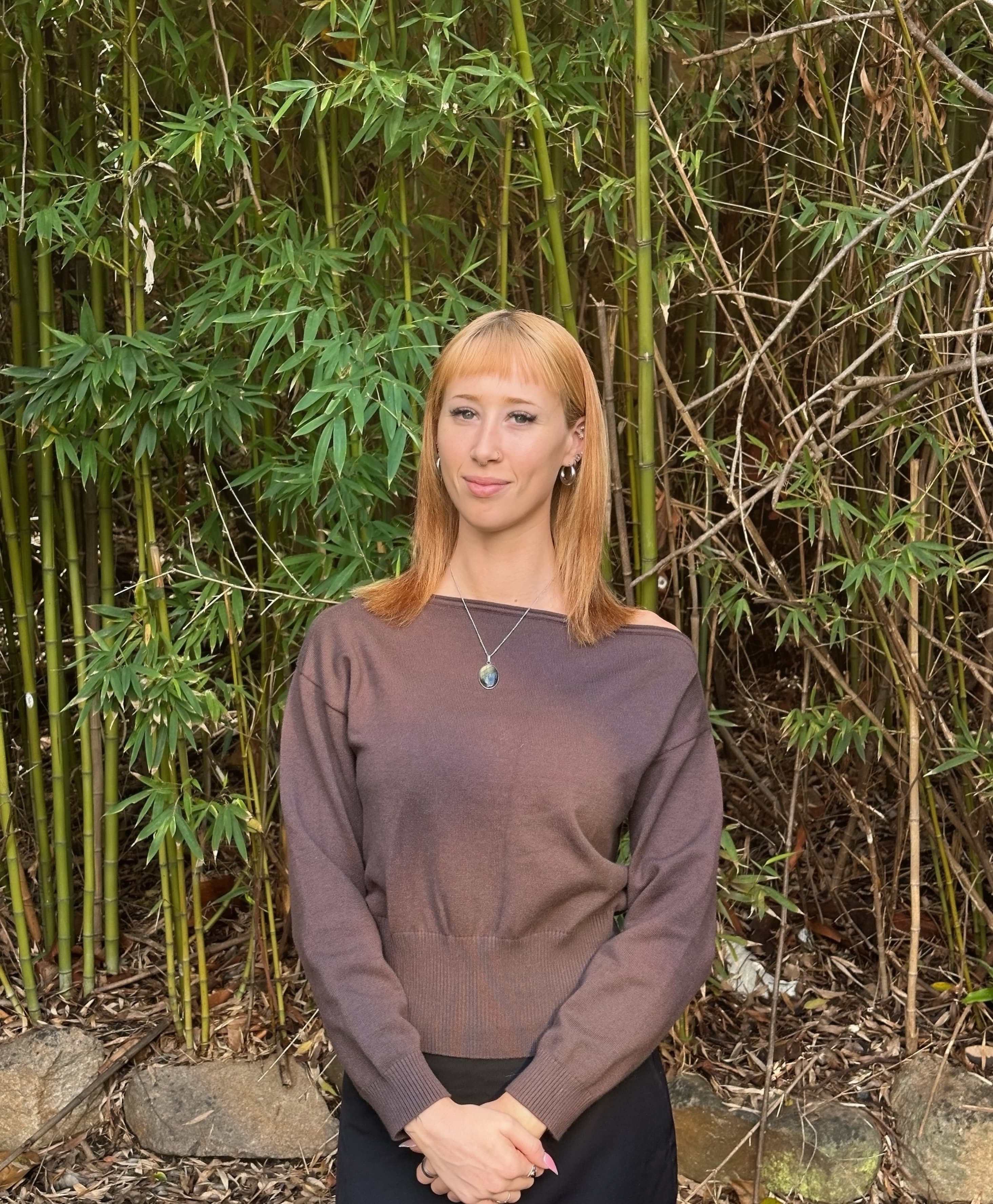 Woman with strawberry blonde hair and bangs wearing a brown sweater against bamboo.